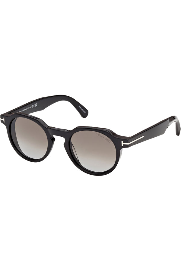 Rotated view of Tom Ford oval sunglasses in gray with black frame and metal T details, shown against a plaid white background.