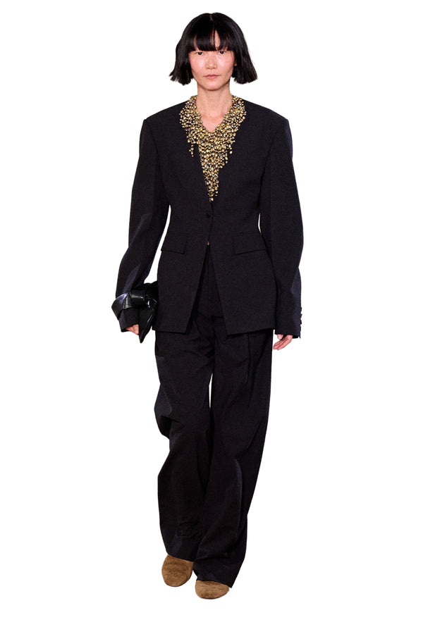 Model wearing Nanushka blazer in black with button, paired with detailed necklace, black trousers and a pair of brown shoes, shown against a plain white background.