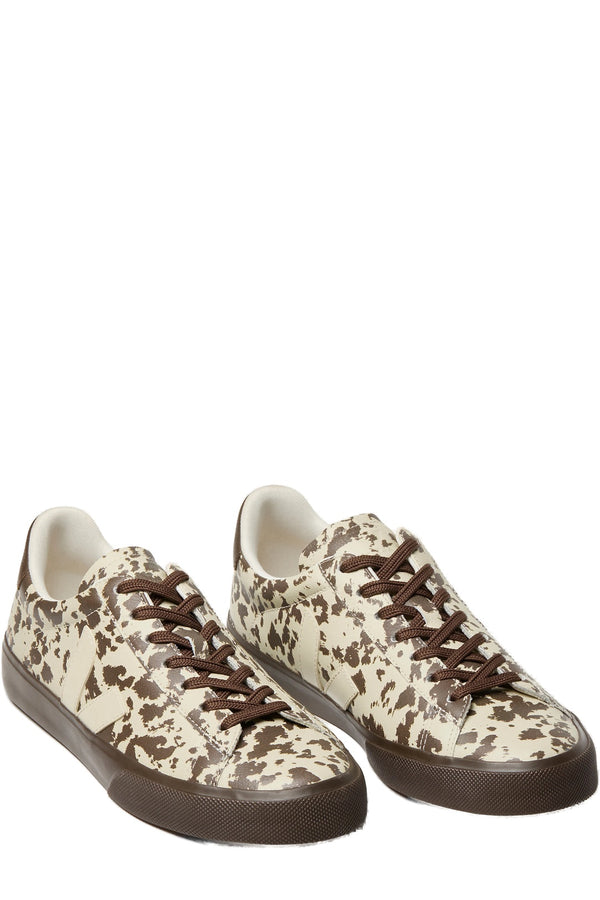A pair of low-top sneakers featuring a brown and cream cow-print pattern, dark brown laces, and dark brown rubber soles, shown against a plain white background.